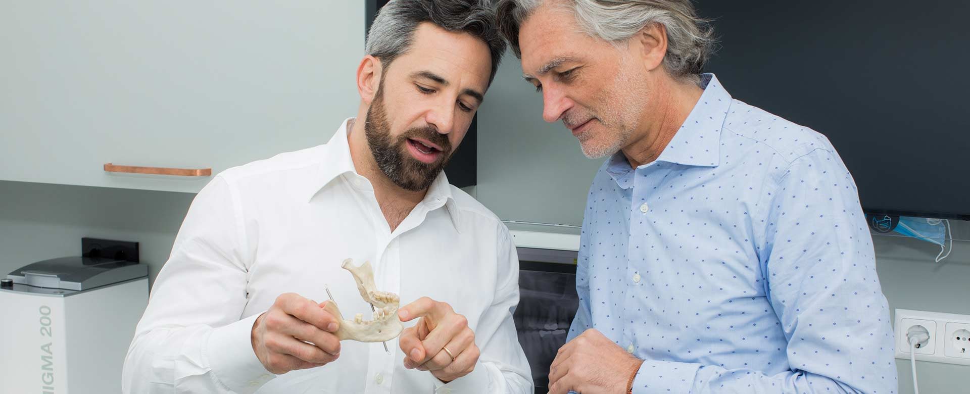 Zahnarzt Dr. Andreas Quidenus erklärt älterem Patienten am Modell eines Unterkiefers den Knochenaufbau
